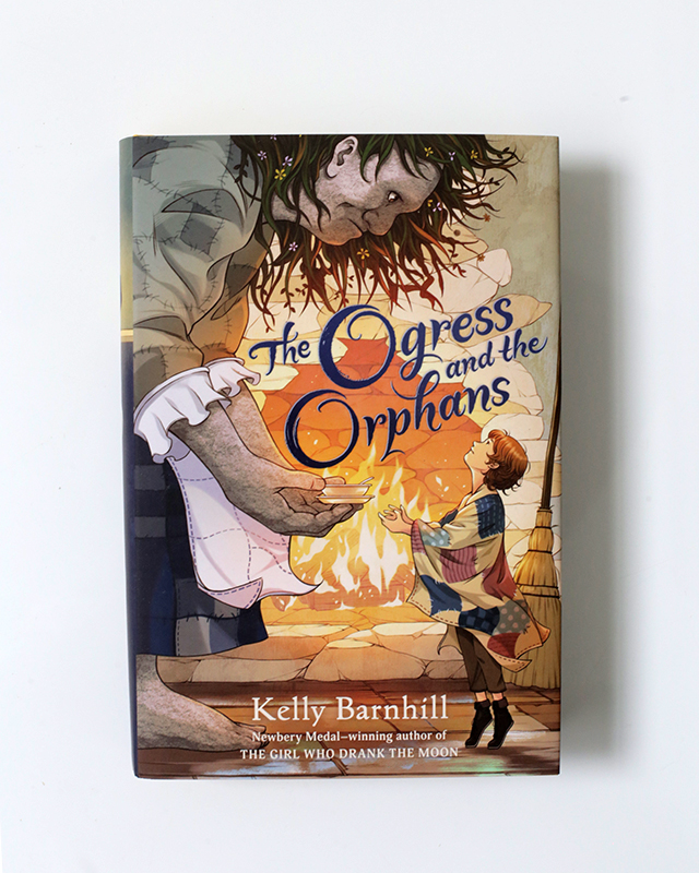 A hard cover middle grade book featuring an giant ogress giving a small child a book in front of a fire.
