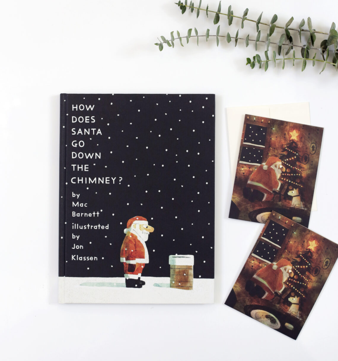 A book lays on a table with two cards that match the book cover. The cover is Santa looking at a snowy chimney.